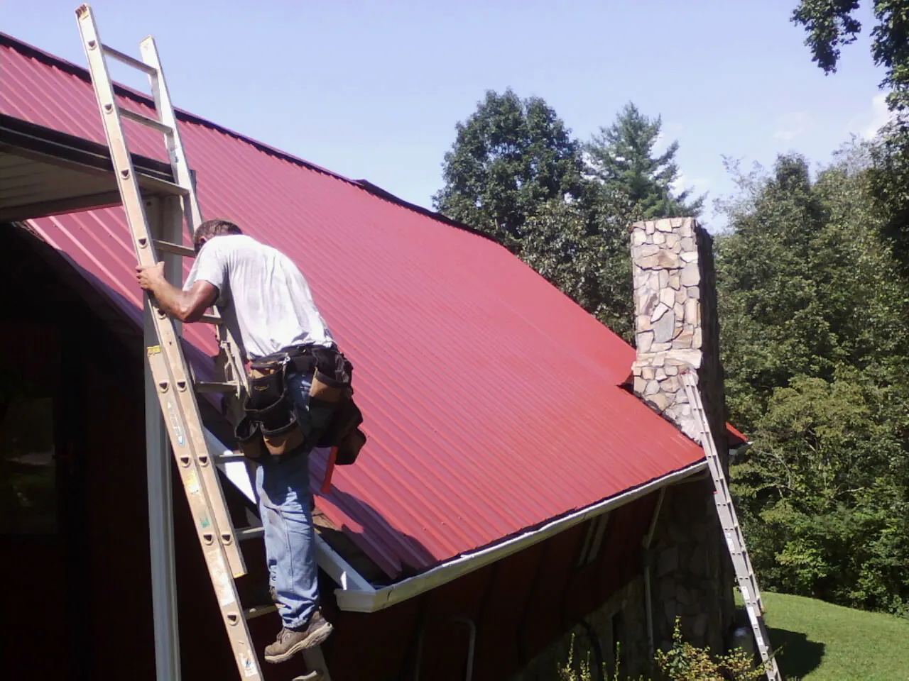 Roofer on a ladder beside a red metal roof and stone chimney — Soffit & Fascia Repair by Roofing Brothers in Breaux Bridge